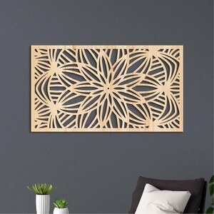 Puede incluir: Una pieza rectangular de arte mural de madera con un diseño floral. La obra de arte está hecha de madera de color claro y presenta un patrón simétrico de flores estilizadas y líneas curvas. La pieza está montada en una pared gris oscuro.