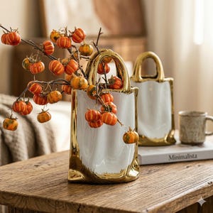 May include: Two white ceramic vases with gold trim, shaped like shopping bags, one filled with branches of small orange pumpkins. A mug and a book are in the background, creating a seasonal display.