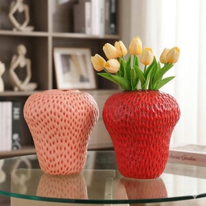 May include: Two ceramic strawberry-shaped vases. One is a light pink color, and the other is bright red. The red vase holds yellow tulips. Both vases have a textured surface with small, evenly spaced indentations.