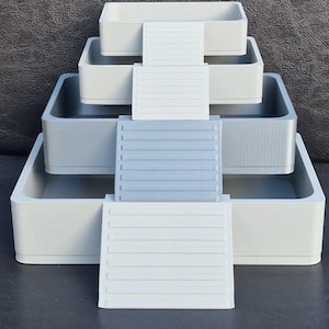Puede incluir: Un organizador de almacenamiento escalonado, de color gris claro, con cuatro bandejas rectangulares de diferentes tamaños. Cada bandeja está conectada por una serie de plataformas horizontales con estrías, creando una apariencia escalonada. Diseñado para guardar objetos pequeños.
