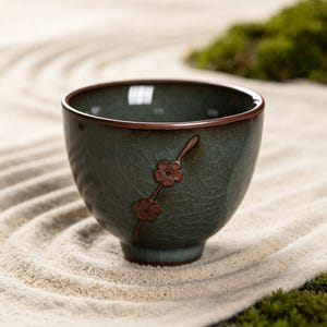 May include: A small, round ceramic tea cup with a dark teal, crackled glaze finish. The exterior features a raised, brown-toned branch motif with two small, five-petaled flowers. The rim of the cup is thin and finished in a dark brown tone.