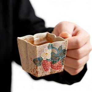 May include: A square ceramic teacup with a strawberry design. The cup has a beige speckled background with a floral and strawberry motif in red, green, and white. The cup is held by a person.