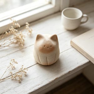 Könnte beinhalten: Eine kleine, abgerundete Keramik-Katzenfigur mit matter, cremeweißer Oberfläche und einem sanft hellbraun gefärbten Gesicht. Die Katze hat einfache, eingravierte Linien für Ohren und Pfoten sowie zwei kleine Punkte als Augen und eine kleine Nase.