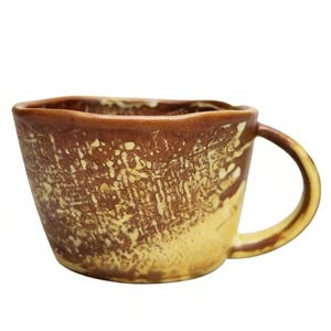 May include: A handmade ceramic mug with a unique, textured surface. The mug features a brown rim and handle, with a gradient of light yellow to brown on the body. The mug has a capacity of approximately 12 ounces.