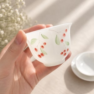 Op de afbeelding: Een witte keramische theekop met een kersendesign. De kop heeft rode kersen en groene bladeren aan de buitenkant. De kop wordt in een hand gehouden, met een bijpassende schotel op de achtergrond.
