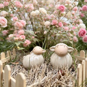 May include: Two ceramic sheep figurines with smooth, glossy off-white bodies and matte beige faces. The sheep are positioned side-by-side, featuring simple, rounded features and small ears, set within a small wooden picket fence enclosure.
