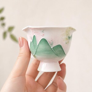 Può includere: Una piccola tazza da tè in ceramica bianca con bordo svasato e base a piedistallo. La tazza è decorata con un motivo a catena montuosa verde e delicati accenti floreali rosa e verdi. La tazza da tè è tenuta in una mano.