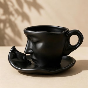 May include: A black ceramic teacup and saucer set. The teacup features a sculpted face design on the side, with a handle. The saucer is a matching black color and has a curved shape. The set is designed for coffee or tea.