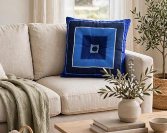 Op de afbeelding: Een vierkant sierkussen met een concentrisch geometrisch patroon in tinten koningsblauw, marineblauw en lichtblauw, met een klein donker cirkelvormig accent in het midden. Het kussen heeft een getuft middelpunt en een gestructureerde rand.