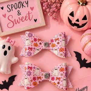 May include: Two hair bows featuring a light pink background with a repeating pattern of white ghosts, orange pumpkins, purple flowers, and pink bows. Each bow has a central clear rhinestone embellishment and glittery pink side accents.