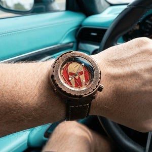 Puede incluir: Un reloj de pulsera de color bronce con un diseño de casco espartano rojo y dorado en la esfera. El reloj tiene una correa de cuero marrón y se lleva en la muñeca. La esfera tiene números y marcas.