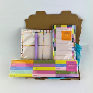 May include: A planner set in a cardboard box. The set includes a priority planner, a pen, a list pad with a magnet, and a keyboard need pad. The items have colourful designs with text that reads "PRIORITY PLANNER" and "THE LIST".