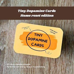 Puede incluir: Una tarjeta rectangular amarilla con esquinas redondeadas, con el texto "Tiny Dopamine Cards" en un círculo negro con fondo naranja. La tarjeta también incluye el texto "Home reset edition" y "If you're stuck, do this."