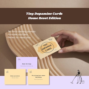 Op de afbeelding: Een set "Tiny Dopamine Cards Home Reset Edition" met schoonmaakopdrachten. De kaarten zijn lichtgeel met zwarte tekst en een cirkelvormig logo. De afbeelding bevat voorbeelden van kaarten met opdrachten als "Reset the fridge" en "Clean one mirror."