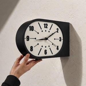 May include: A wall-mounted clock with a matte black casing and a white face. The clock features black Arabic numerals for 2, 3, 4, 6, 8, 9, 10, and 12, with black line markers and dots for the remaining positions. It has black hour, minute, and second hands.