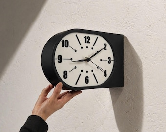 May include: A wall-mounted clock with a matte black casing and a white face. The clock features black Arabic numerals for 2, 3, 4, 6, 8, 9, 10, and 12, with black line markers and dots for the remaining positions. It has black hour, minute, and second hands.