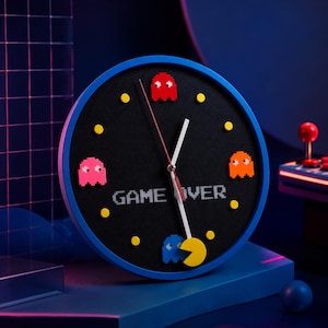 Puede incluir: Un reloj de pared redondo con marco azul y esfera negra que presenta figuras de fantasmas pixeladas en rojo, rosa, naranja y azul. La esfera muestra el texto GAME OVER en fuente pixelada blanca, con puntos amarillos y un personaje amarillo.