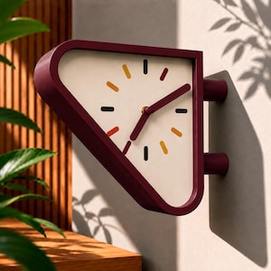 Puede incluir: Un reloj de pared triangular con un marco de color granate intenso y una esfera de color crema. El reloj presenta marcadores de hora negros, amarillos y naranjas, junto con dos manecillas granates. Se fija a la pared mediante dos soportes.