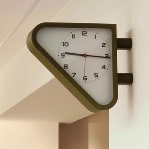 Könnte beinhalten: Eine Wanduhr mit einem dreieckigen Rahmen in gedämpftem Olivgrün. Das Zifferblatt ist weiß mit schwarzen Ziffern von 2 bis 12, außer der 1, und verfügt über schwarze Stunden- und Minutenzeiger sowie einen dünnen roten Sekundenzeiger.