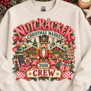 May include: A beige crewneck sweatshirt featuring a graphic of a nutcracker standing in a Christmas market. The text reads NUTCRACKER CHRISTMAS MARKET, 2026 CREW, 'TIS THE SEASON, and GOOD THINGS HAPPEN HERE. The design includes red bows, candy canes, and gifts.