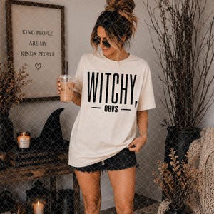 May include: A cream-colored short-sleeve t-shirt featuring the black text WITCHY, OBVS printed on the front. The shirt has a crew neckline and a relaxed fit, paired with black denim shorts. The design is centered on the chest in a bold font.