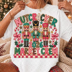 Può includere: Una t-shirt bianca con una grafica di tre schiaccianoci circondati da bastoncini di zucchero, stelle e foglie di agrifoglio. Il testo recita NUTCRACKER 2026 MARKET CREW. Il design utilizza uno stile patchwork con motivi rossi, verdi e leopardati.