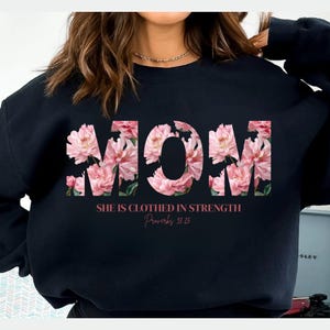 May include: Black sweatshirt featuring the word "MOM" in a floral design with pink and white flowers. The text "SHE IS CLOTHED IN STRENGTH Proverbs 31:25" is printed below the word "MOM".