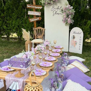 May include: A bridal shower picnic setup with a wooden table, decorated with purple and white accents. The table is set with plates, napkins, and glasses. A cake, gifts, and pillows are arranged on the grass. A sign says "Bridal Shower".