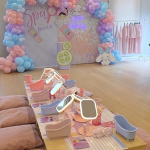Puede incluir: Una fiesta de spa con una mesa con bañeras en miniatura, espejos y productos de belleza. La decoración incluye un arco de globos en colores pastel, un telón de fondo con lentejuelas y un personaje de dibujos animados. Las palabras "Spa Party" son visibles.