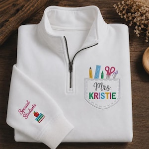 Op de afbeelding: Witte quarter-zip sweatshirt met een zwarte rits. De borstzak heeft een ontwerp met schoolspullen en de naam "Mrs. KRISTIE". De linkermouw heeft de woorden "Special Students" en een appel- en boekenontwerp.