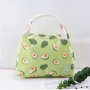 May include: A light green lunch bag featuring a repeating pattern of avocado halves with dark green skins and orange pits. The bag has a top zip closure, two beige woven fabric handles, and a small square label with an avocado icon.