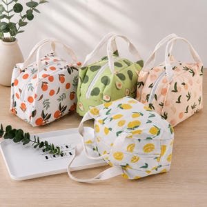 May include: Four rectangular lunch bags with top handles and zip closures. Each bag features a distinct fruit or vegetable pattern: one white with oranges, one green with avocados, one peach-coloured with peaches, and one white with yellow lemons.