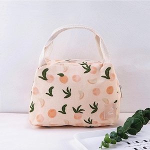 May include: A light peach-coloured lunch bag featuring a repeating pattern of orange fruit and dark green leaves. The bag has a top handle made of cream-coloured woven fabric and a small square label on the front with the word Peach printed on it.
