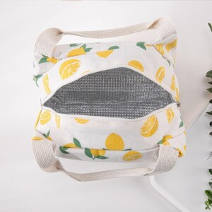 May include: A white insulated lunch bag featuring a repeating pattern of yellow lemon slices and green leaves. The bag has two beige woven handles and a silver metallic thermal lining visible through the open zippered top closure.