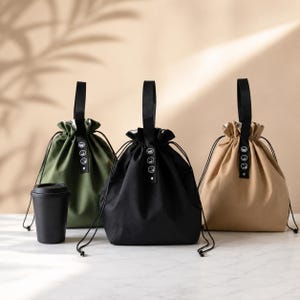 May include: Three drawstring bags in olive green, black, and beige, each featuring a black top handle with a vertical strip containing three white circular smiley face icons. A solid black lidded cup stands next to the olive green bag.