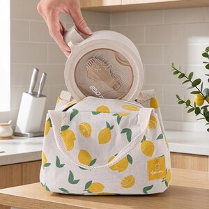 May include: A beige fabric lunch bag featuring a repeating pattern of yellow lemons and green leaves, with two sturdy handles. A round, beige 850 ml lunch box with the text "LUNCH BOX" printed on the lid is being placed inside the bag.