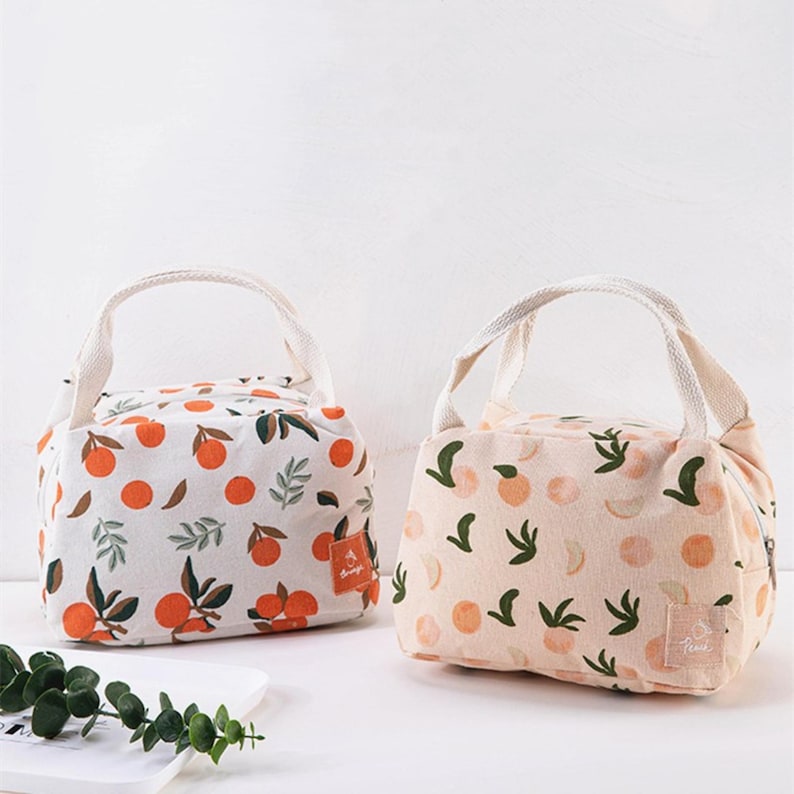 May include: Two rectangular lunch bags with top handles and zip closures. The left bag is white with an orange fruit and green leaf pattern. The right bag is light peach with a peach fruit and green leaf pattern, featuring a small label reading Peach.