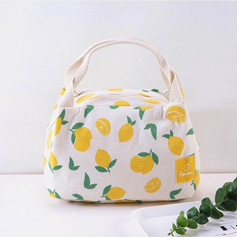May include: A cream-coloured fabric lunch bag featuring a repeating pattern of yellow lemons and green leaves. The bag has two cream-coloured woven handles and a small square yellow patch on the lower right side with the word lemon printed on it.