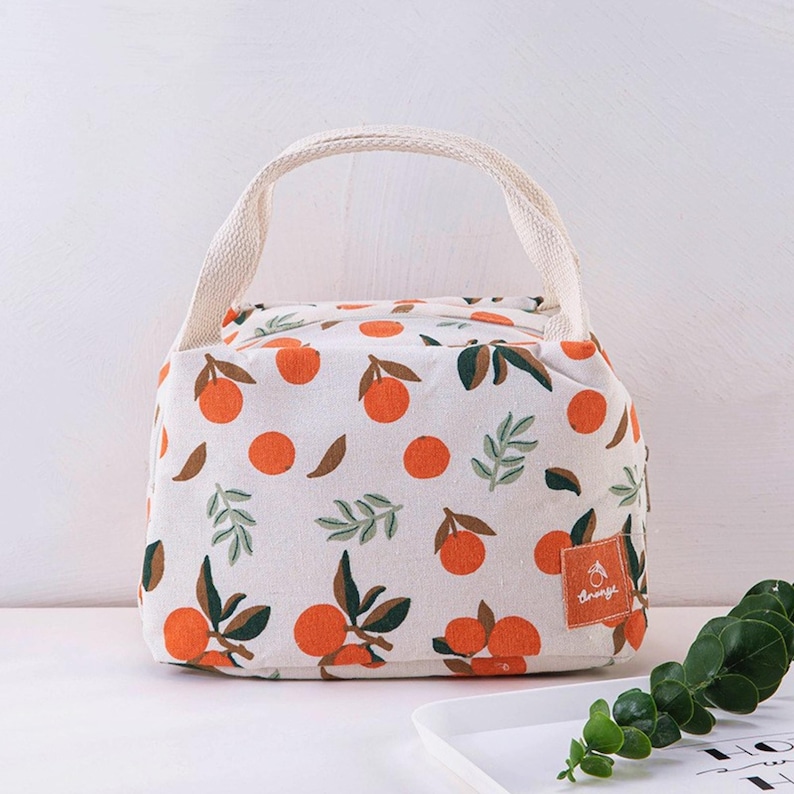 May include: A cream-coloured lunch bag featuring a repeating pattern of bright orange citrus fruits and dark green leaves. The bag has two woven fabric handles and a small rectangular orange label on the bottom right corner with white text.