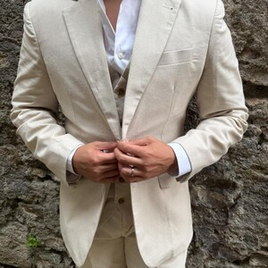 May include: A light beige linen suit, consisting of a blazer, vest, and trousers, paired with a white dress shirt. The suit is tailored and appears to be made of a lightweight fabric. The person is buttoning the blazer. The background is a stone wall.