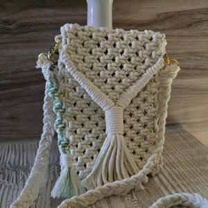 Puede incluir: Bolso cruzado de macramé color crema con una correa trenzada y detalles de borlas. El bolso presenta un diseño tejido y herrajes dorados. Un pequeño jarrón blanco con flores moradas está en el fondo.