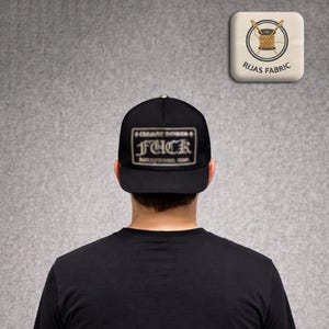 Pode incluir: Boné de beisebol preto com aba reta. O boné apresenta um patch retangular com a palavra "FUCK" em letras em negrito e o texto "Chrome Hearts Hollywood, CA". O fundo é uma parede cinza.