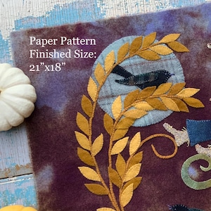 May include: A fabric applique wall hanging featuring a scarecrow in a plaid hat, blue overalls, and a checkered shirt sitting on a large pumpkin. The scene includes a black cat, two black birds, a golden vine, and a moon. Text reads: Paper Pattern Finished Size: 21"x18".