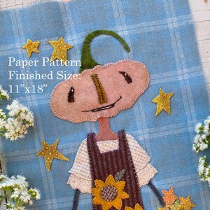 May include: A fabric applique wall hanging on a blue plaid background featuring a pumpkin-headed figure wearing a brown striped apron with sunflowers, holding a basket and bucket of stars. Text reads: Paper Pattern Finished Size: 11"x18".