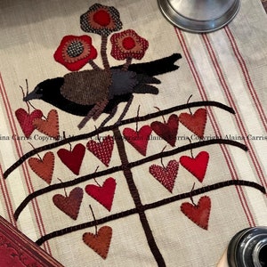 Puede incluir: Un pájaro negro posado en una rama marrón con corazones de fieltro rojo colgando de ella. El fondo es una tela de color crema con rayas rojas y blancas. El texto "Copyright Alaina Carris" es visible en la parte inferior de la imagen.