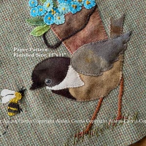 May include: A felt applique of a bird carrying a pot of blue forget-me-not flowers. The bird is brown, gray, and white. The background is a green tweed fabric. The text "Paper Pattern Finished Size: 1"x11"" is visible in the image.