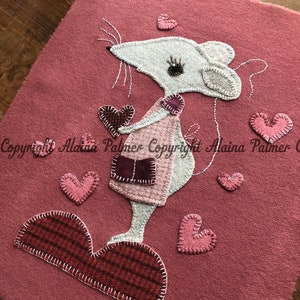 May include: A white felt mouse holding a red felt heart, standing on a red felt heart with pink felt hearts scattered around it. The background is a pink felt.