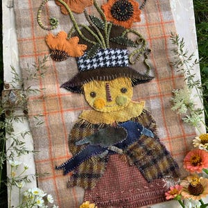 Puede incluir: Un tapiz textil que muestra una figura de espantapájaros con cara amarilla, que lleva un sombrero marrón con una banda de pata de gallo en blanco y negro. La figura sostiene un cuervo azul oscuro sobre un fondo de tela de lana a cuadros.