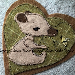 Puede incluir: Un tapiz de pared en forma de corazón de fieltro con un oso marrón y una abeja. El oso es marrón con una cara marrón claro y los ojos cerrados. La abeja es amarilla y negra con un cuerpo marrón. El fondo es una tela de cuadros verdes.