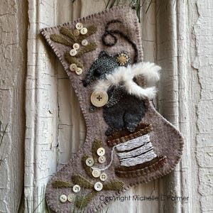 May include: A brown felt stocking with a gray mouse holding a spool of white thread. The mouse is wearing a gray hat with a button. The stocking is decorated with green leaves and white buttons.